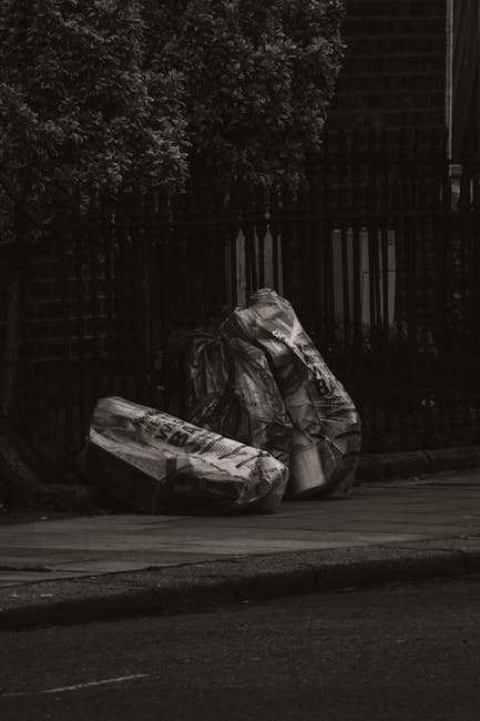 A black and white photograph showing three large, crumpled black plastic garbage bags piled against a wooden fence on a paved sidewalk. The bags are weathered and appear to be filled with household waste, with some sections bulging and torn. In the background, there is a tall wooden fence with vertical slats, partially obscured by leafy trees and shrubs. The environment suggests an outdoor urban or residential area, with the bags waiting for collection. The scene highlights the need for proper rubbish disposal, which Rubbish Removal Bermondsey offers through private waste handling and alternative collection services, maintaining a neat and clean streetscape.
