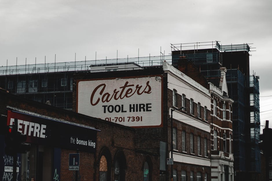 A multi-story building with a prominent white sign for 'Carters Tool Hire' in dark red and black lettering, mounted on the brick facade of the structure. The building appears to be part of a commercial area, with scaffolding and construction work visible on the roof and upper sections, indicating ongoing renovation or expansion. To the left, a storefront with signage that includes the words 'the bonus king' is partially visible. The scene is set outdoors during overcast weather, with a cloudy grey sky overhead. The building features a combination of red brick and white painted trim around the windows and cornices, with large rectangular windows aligned symmetrically across the facade. The overall scene suggests urban commercial activity, and the image captures the building’s exterior from a slightly angled perspective, emphasizing its height and the construction activity in progress, relevant to contexts of property renovation, development, or alternative waste handling associated with building works. Rubbish Removal Bermondsey handles waste from such renovation projects, supporting safe and efficient clearance of construction debris.