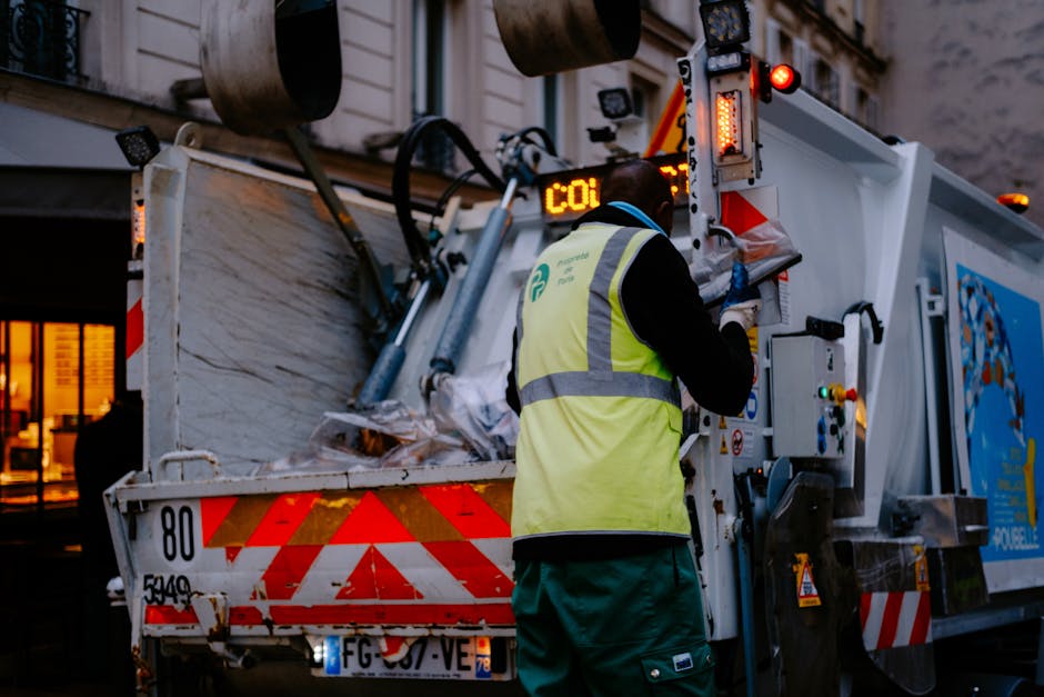 A waste collection worker wearing a high-visibility yellow vest and gloves operates a large urban rubbish truck on a street at dusk. The truck is equipped with hydraulic lifting mechanisms and has a partially open rear compartment loaded with various waste materials, including paper, plastic, and other refuse. The vehicle features a white body with red reflective warning stripes along its edges and an illuminated electronic display on top. The background shows a building with lit windows, creating a dimly lit urban atmosphere suitable for evening waste collection, demonstrating the typical scene of private rubbish removal with a focus on efficient on-site waste handling and disposal, aligned with independent rubbish removal services.