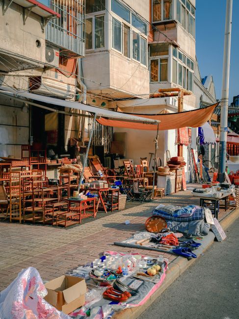 The image depicts a street-side outdoor area adjacent to a multi-storey building with large glass windows and concrete walls. In the foreground, various items are laid out on the pavement, including plastic bottles, cardboard boxes, and miscellaneous household objects, suggesting a cluttered display or clearance sale. To the right, a collection of wooden chairs and tables are stacked and arranged haphazardly beneath a beige and orange sunshade canopy, providing partial shade to the items. Behind the outdoor setup, there are additional furniture pieces, some covered with fabric or plastic, and a few textiles hanging vertically, which may be drying or for sale. The building's exterior features multiple balconies with metal railings, air conditioning units, and visible piping, creating an urban environment. The overall scene is lit naturally, with daylight casting shadows, and the image fits within a context of private waste handling or on-site clearance that might be performed by a rubbish removal service, such as Rubbish Removal Bermondsey, to manage clutter from market or street trader activities.