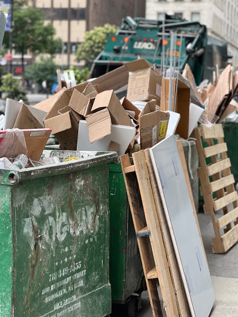 A large green waste bin filled with assorted cardboard boxes, some flattened and others in their original shape, alongside a wooden pallet leaning against the side. The cardboard comprises various sizes and shades of brown, with some boxes featuring labels or printed text. In the background, there is a green waste collection vehicle with visible branding and orange lights, parked on an urban street with surrounding buildings and trees. The scene suggests a recent or ongoing rubbish removal process, with materials ready for disposal or recycling, typical of private waste handling services offered by companies such as Rubbish Removal Bermondsey. The environment is outdoor, with natural daylight illuminating the items, and the overall setting indicates an on-site clearance of waste materials related to domestic or commercial refuse collection in a central London location.