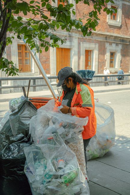 A woman wearing a dark baseball cap, a black face mask, and an orange high-visibility jacket with reflective yellow-green stripes is standing on a city sidewalk, surrounded by several large black and clear plastic rubbish bags filled with various waste items. She appears to be sorting or inspecting the contents of a transparent bag, which contains plastic bottles, food containers, and other light debris. In the background, there is a brick building with stone window frames and a metal railing along the sidewalk, with a tree branch with green leaves partially framing the upper part of the image. The scene suggests an on-site or private waste collection activity, likely organized by a rubbish removal service such as Rubbish Removal Bermondsey, in an urban environment, with natural daylight providing clear visibility of the scene's details and textures.