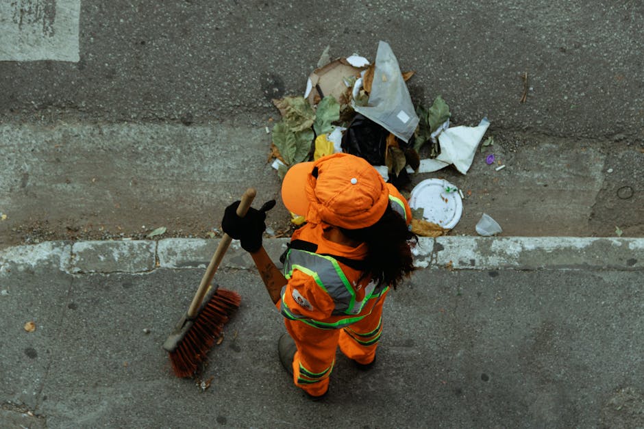 A large green waste bin filled with assorted cardboard boxes, some flattened and others in their original shape, alongside a wooden pallet leaning against the side. The cardboard comprises various sizes and shades of brown, with some boxes featuring labels or printed text. In the background, there is a green waste collection vehicle with visible branding and orange lights, parked on an urban street with surrounding buildings and trees. The scene suggests a recent or ongoing rubbish removal process, with materials ready for disposal or recycling, typical of private waste handling services offered by companies such as Rubbish Removal Bermondsey. The environment is outdoor, with natural daylight illuminating the items, and the overall setting indicates an on-site clearance of waste materials related to domestic or commercial refuse collection in a central London location.