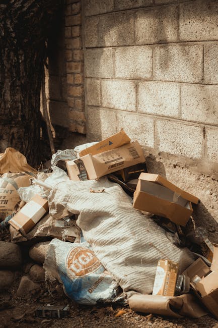 A pile of discarded cardboard boxes and paper waste piled against a rough, exposed brick and concrete wall outdoors. The boxes vary in size, some with flaps open revealing empty interiors, while others are crushed or partially collapsed. Several large bags, possibly made of woven polypropylene, contain additional waste and are partially covered by a fabric, with visible creases and wear. The scene is illuminated by natural light, casting soft shadows on the debris and the surrounding ground, which consists of dirt and small stones. The presence of this waste suggests a site designated for rubbish collection or an area awaiting removal by a rubbish clearance service such as Rubbish Removal Bermondsey, emphasizing the context of private or alternative waste handling rather than municipal collection.