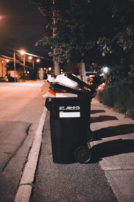 A black wheelie bin labeled with the name 'St. John's' is placed on a sidewalk beside a curb on a quiet residential street at night. The bin is partially filled with waste items, including cardboard and black plastic bags, which are visible at the open top. The bin's surface appears slightly reflective, and the legal lid is propped open, revealing the contents stacked inside. The surrounding environment includes a row of trees and shrubbery on the right side of the pavement, with some branches casting shadows on the ground. In the background, streetlights illuminate the street with a warm orange glow, and faint outlines of parked cars and residential houses are visible under the nighttime sky. The scene suggests an instance of individual waste disposal, where private or alternative rubbish collection methods such as personal bin use might be occurring, which is relevant to proper waste management practices emphasized by rubbish removal services like Rubbish Removal Bermondsey.