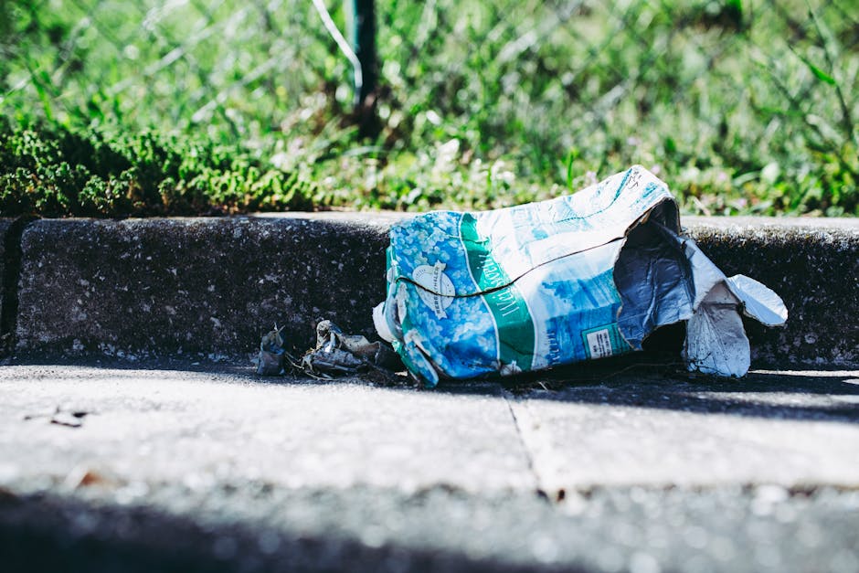 A pile of discarded cardboard boxes and paper waste piled against a rough, exposed brick and concrete wall outdoors. The boxes vary in size, some with flaps open revealing empty interiors, while others are crushed or partially collapsed. Several large bags, possibly made of woven polypropylene, contain additional waste and are partially covered by a fabric, with visible creases and wear. The scene is illuminated by natural light, casting soft shadows on the debris and the surrounding ground, which consists of dirt and small stones. The presence of this waste suggests a site designated for rubbish collection or an area awaiting removal by a rubbish clearance service such as Rubbish Removal Bermondsey, emphasizing the context of private or alternative waste handling rather than municipal collection.